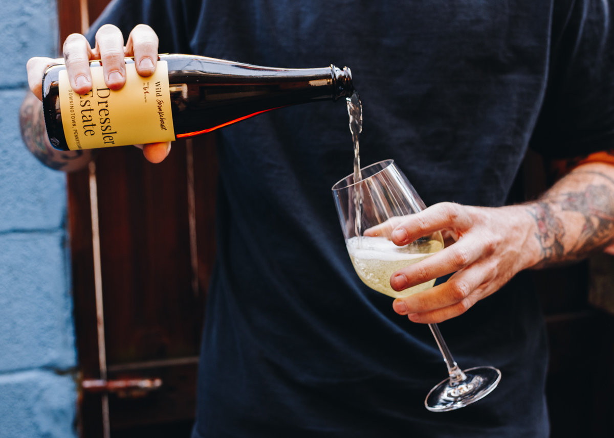 A 500 mL bottle of Dressler Estate's Wild Smokehouse cider being poured by a man into a glass.