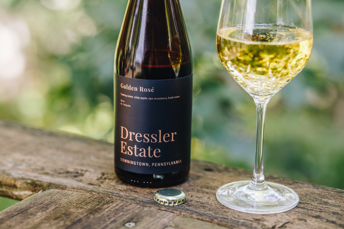 A 500 mL bottle of Dressler Estate's Golden Rose cider with a filled glass on a railing with blurred trees in the background.