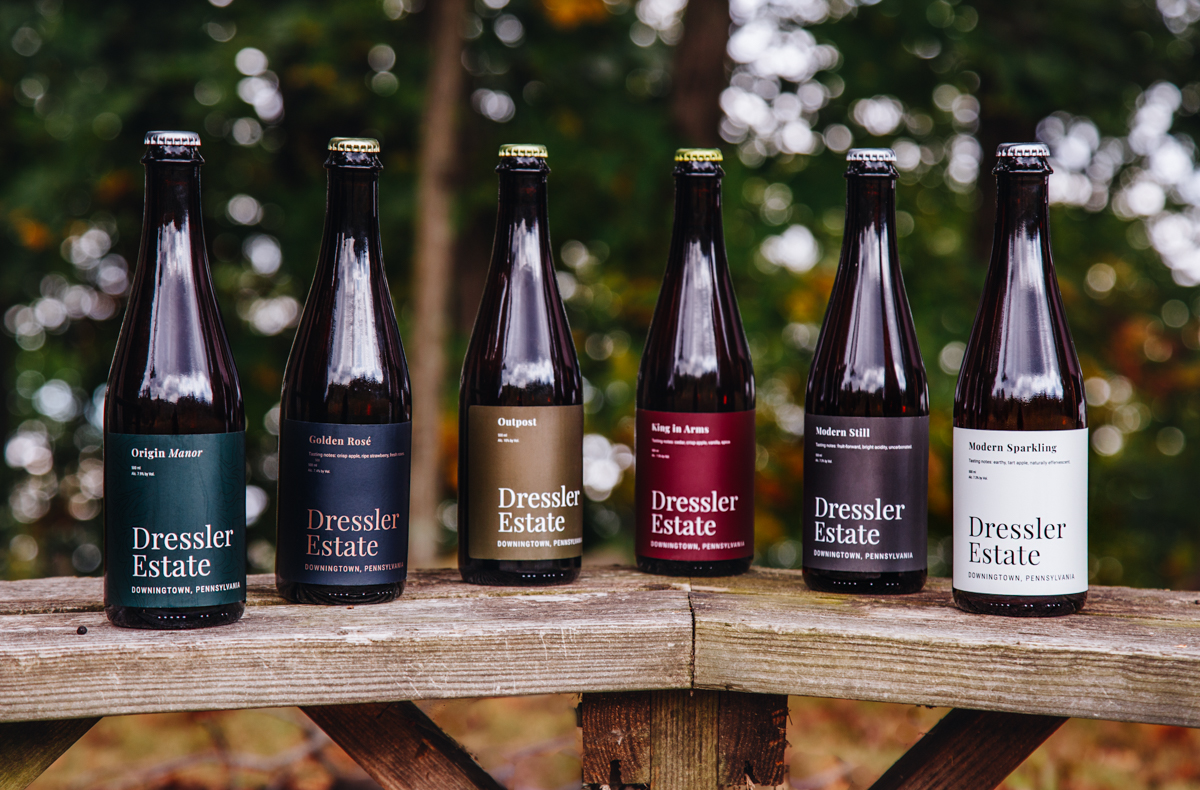 A collection of Dressler Estate cider bottles placed on a bench with trees in the background.