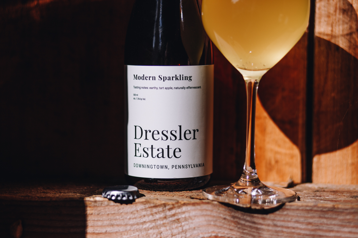 A 500 mL bottle of Dressler Estate's Modern Sparkling cider with a filled glass on a ledge in a barn.