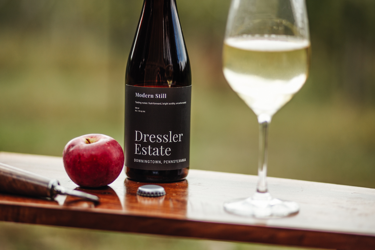 A 500 mL bottle of Dressler Estate's Modern Still cider with a filled glass, apple and bottle opener on a table.
